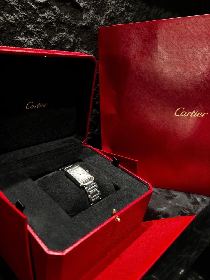 Reloj rectangular de esfera clara y números romanos, dentro de un estuche rojo abierto de Cartier, con la bolsa roja al lado.