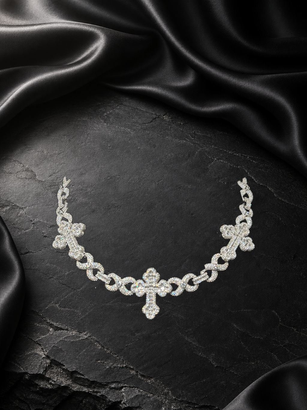 Cadena de eslabones en metal blanco con tres cruces de piedras claras, sobre piedra oscura y satén negro.