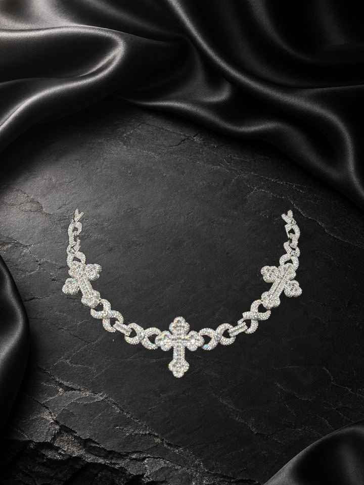 Cadena de eslabones en metal blanco con tres cruces de piedras claras, sobre piedra oscura y satén negro.