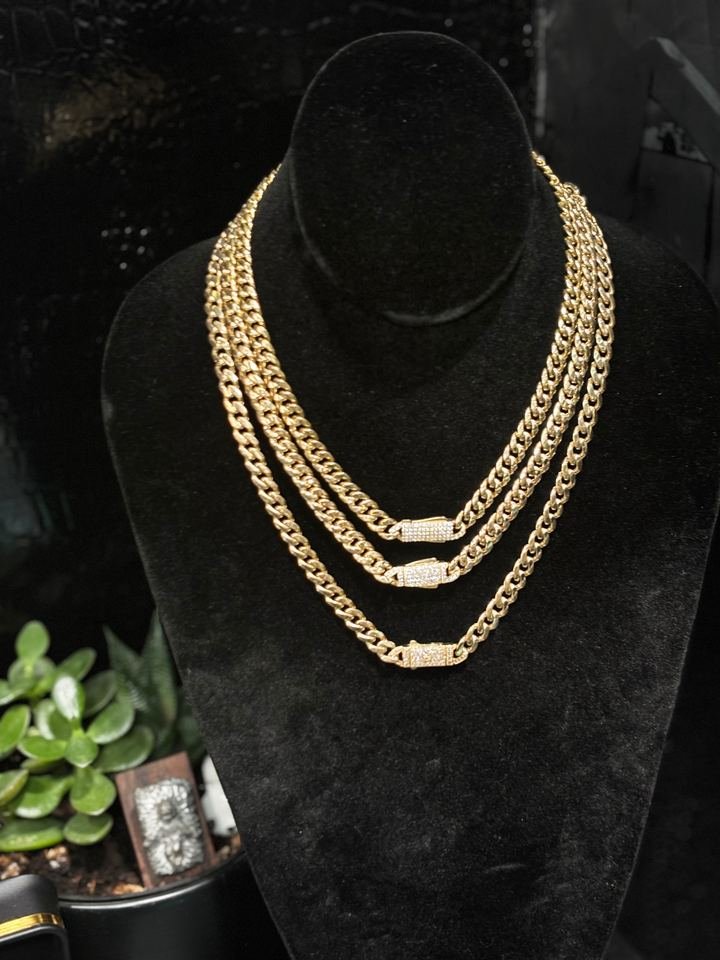 Tres cadenas Miami cubanas de oro amarillo, de largos distintos, colgadas en un busto de terciopelo negro, cada una con el cierre de caja cubierto de piedras blancas.