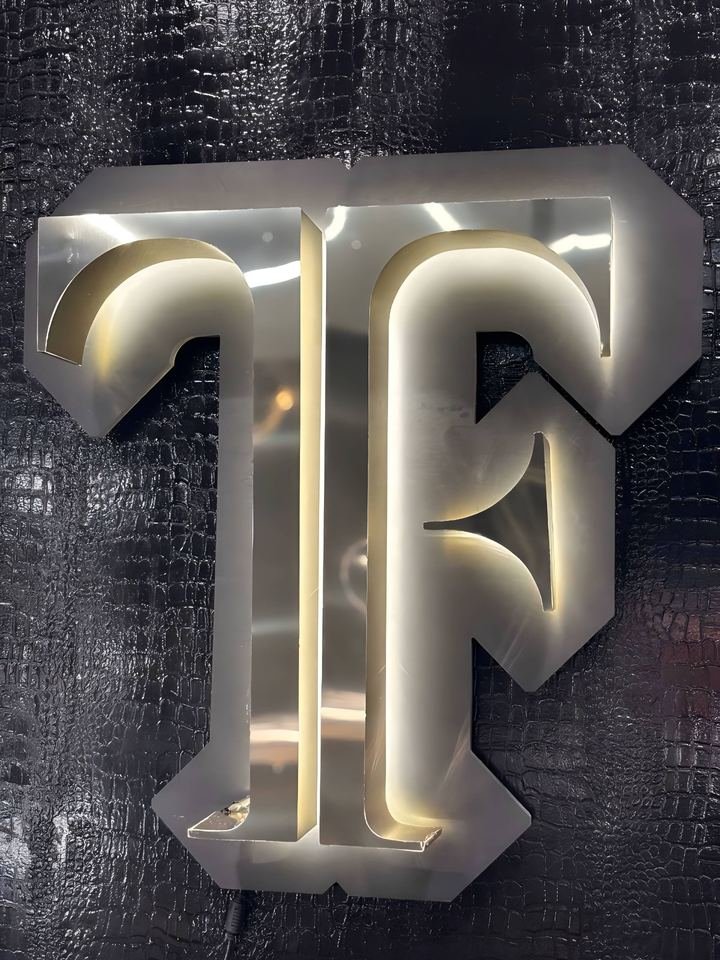 Monograma TF en metal pulido, retroiluminado con luz cálida sobre la pared negra de la tienda.