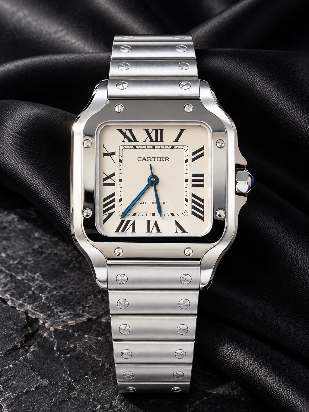 Reloj cuadrado de acero con esfera blanca y números romanos, sobre satén negro y piedra oscura.