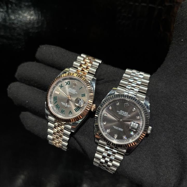 Dos relojes Rolex sostenidos en una mano con guante negro, uno bicolor de acero y oro rosa con números romanos, otro de acero con índices de diamante; los dos con ventanilla de fecha.