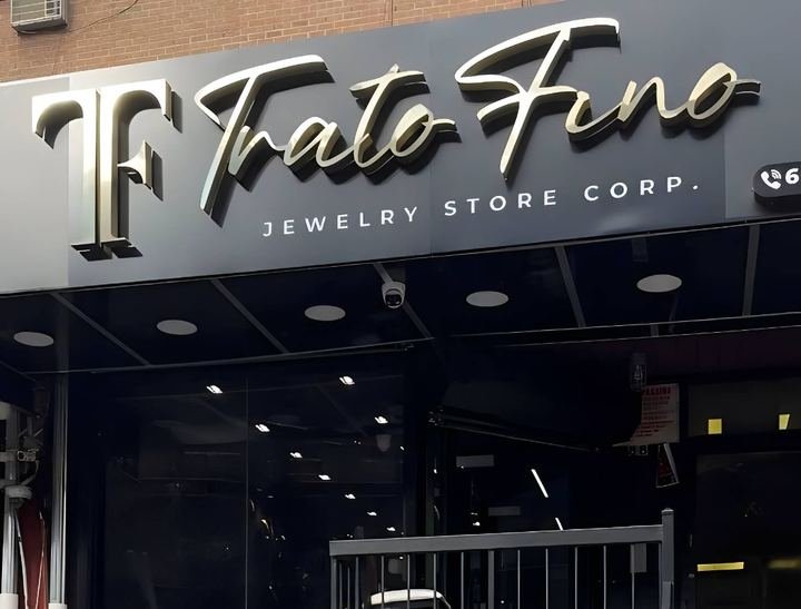 Rótulo exterior de la tienda: el monograma TF y el nombre Trato Fino en letras doradas en relieve sobre una plancha oscura, con Jewelry Store Corp. en mayúsculas al pie. Debajo, el frente de cristal y las luces del portal.