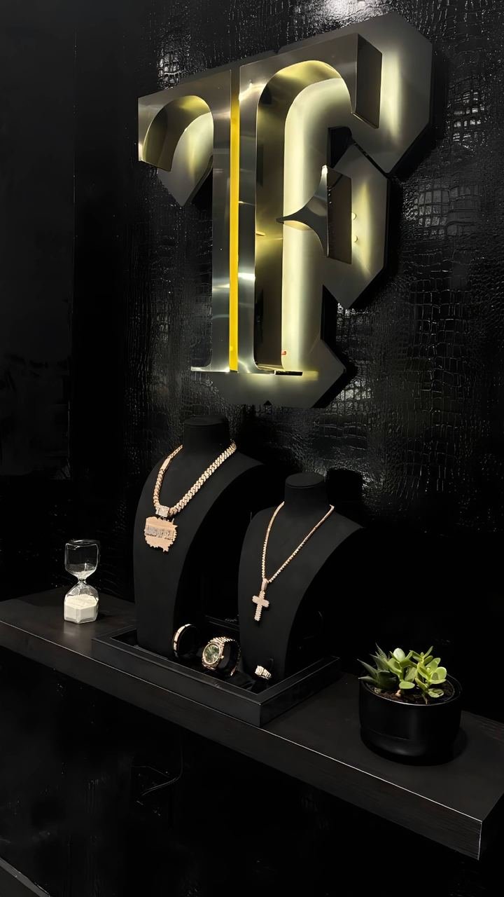 Rótulo TF retroiluminado sobre la pared negra, con dos bustos de terciopelo con cadenas y dijes en la repisa de abajo.